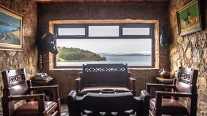 Una habitación rústica con un ventanal grande con vista a un lago, sillas de madera, una mesita y paredes de piedra.