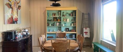 A dining area with a wooden table and chairs, a coffee station, a bookshelf with various items, and a window with curtains.