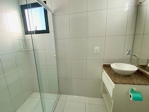 A bathroom with white tiles, a glass shower enclosure, a sink with a marble countertop, and a small window. - Edifício Morada do Imperador (Praia Grande)