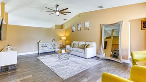 Un salón amplio con suelo de madera, un sofá blanco, un sillón amarillo y una mesa de centro de vidrio.