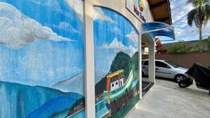 A mural of a boat on a beach with clouds, a car, and a palm tree in the background. - Pousada Mar Suites (Ubatuba)