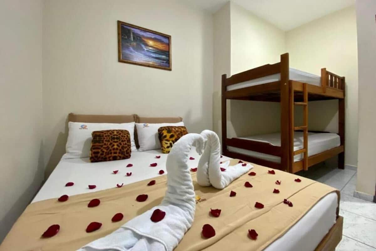 Une chambre d’hôtel avec un lit décoré de pétales de rose et une serviette en forme de cœur. Il y a un lit superposé sur la droite et un tableau au mur.