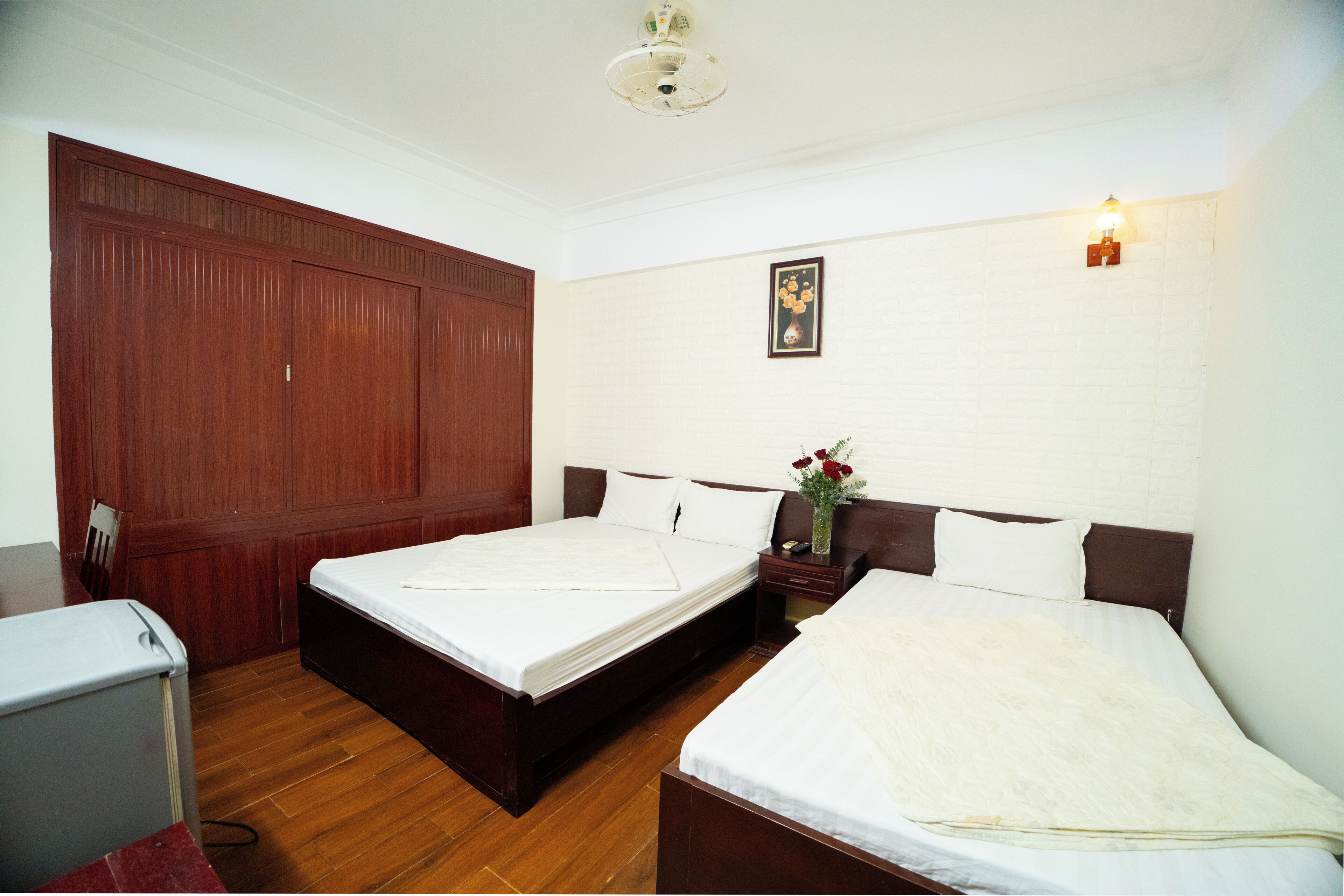 A room with two beds, a wooden wardrobe, a desk, and a small table with a vase of flowers.
