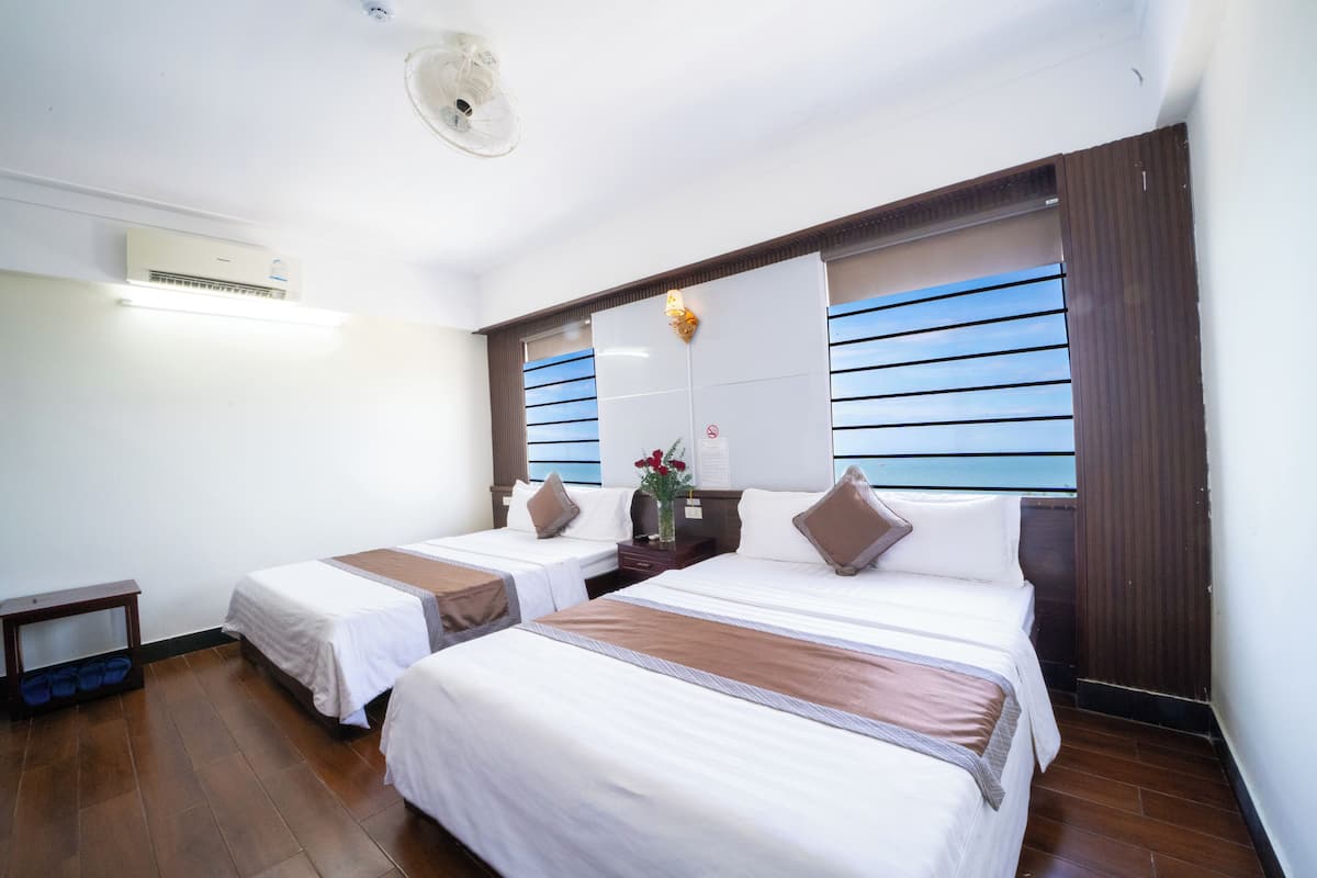 A hotel room with two beds, a ceiling fan, an air conditioning unit, a small table, and a window with blinds.