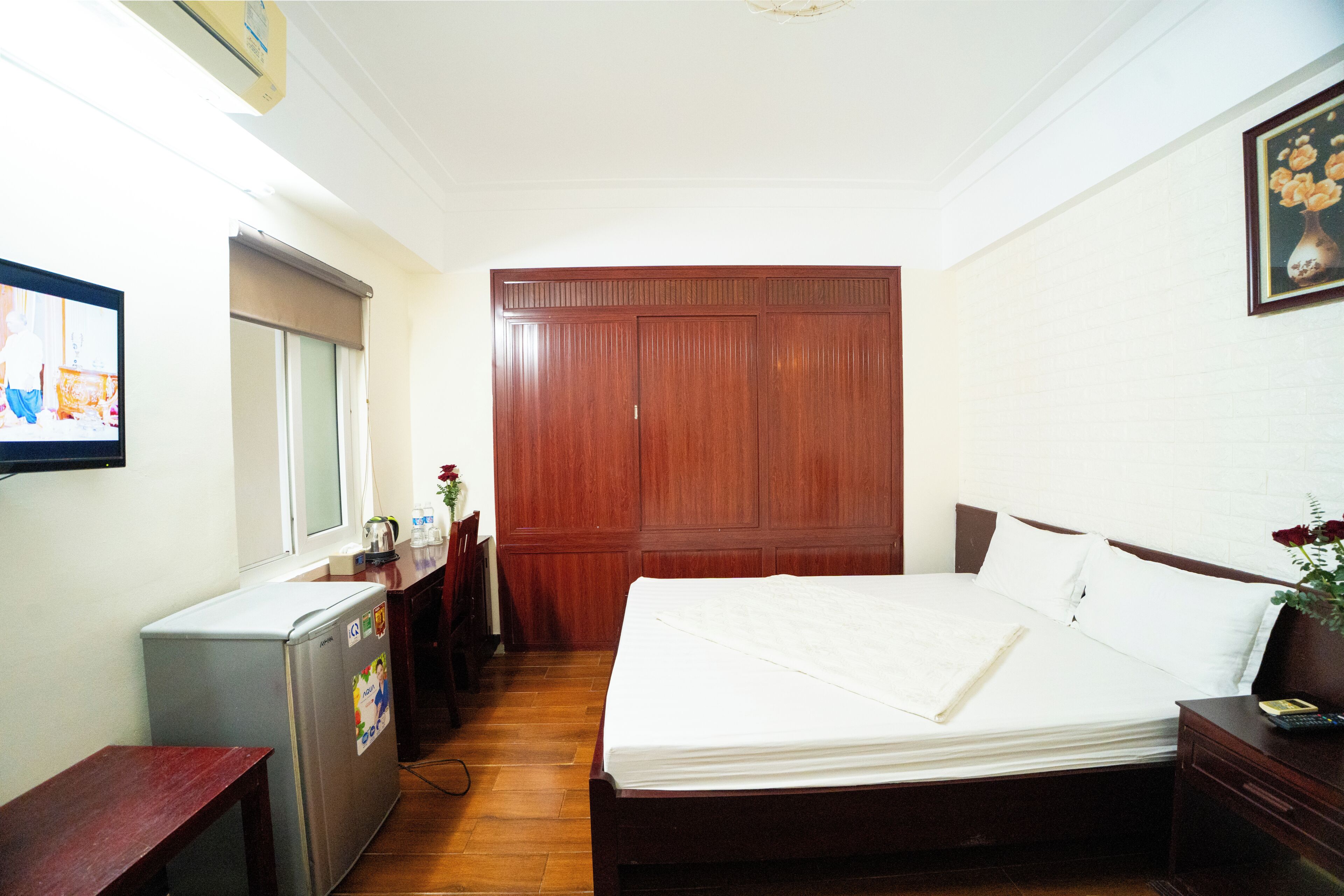 Un dormitorio con cama, armario de madera, refrigerador pequeño, mesita de noche y televisor instalado en la pared.