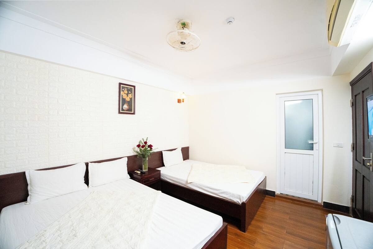A hotel room with two beds, a wooden floor, a white brick wall, a door with frosted glass, and a ceiling light fixture.