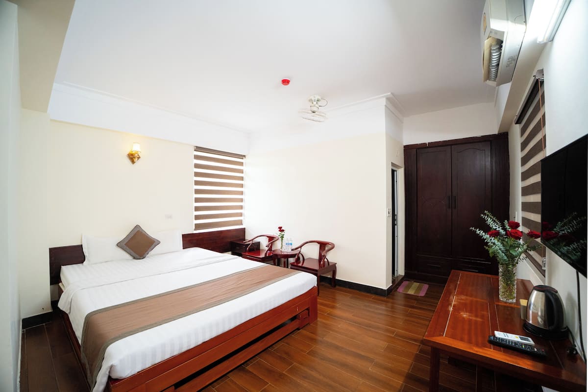 A hotel room with a bed, a desk, a chair, a vase with flowers, a television, and a wardrobe.