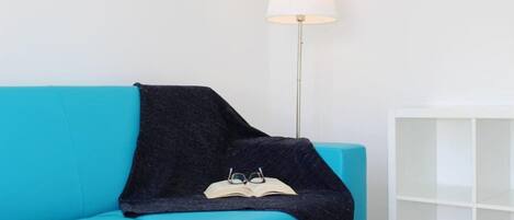 Un canapé de couleur bleu-vert avec un plaid foncé et une paire de lunettes posées dessus. Une lampe de sol blanche se trouve à côté du canapé.