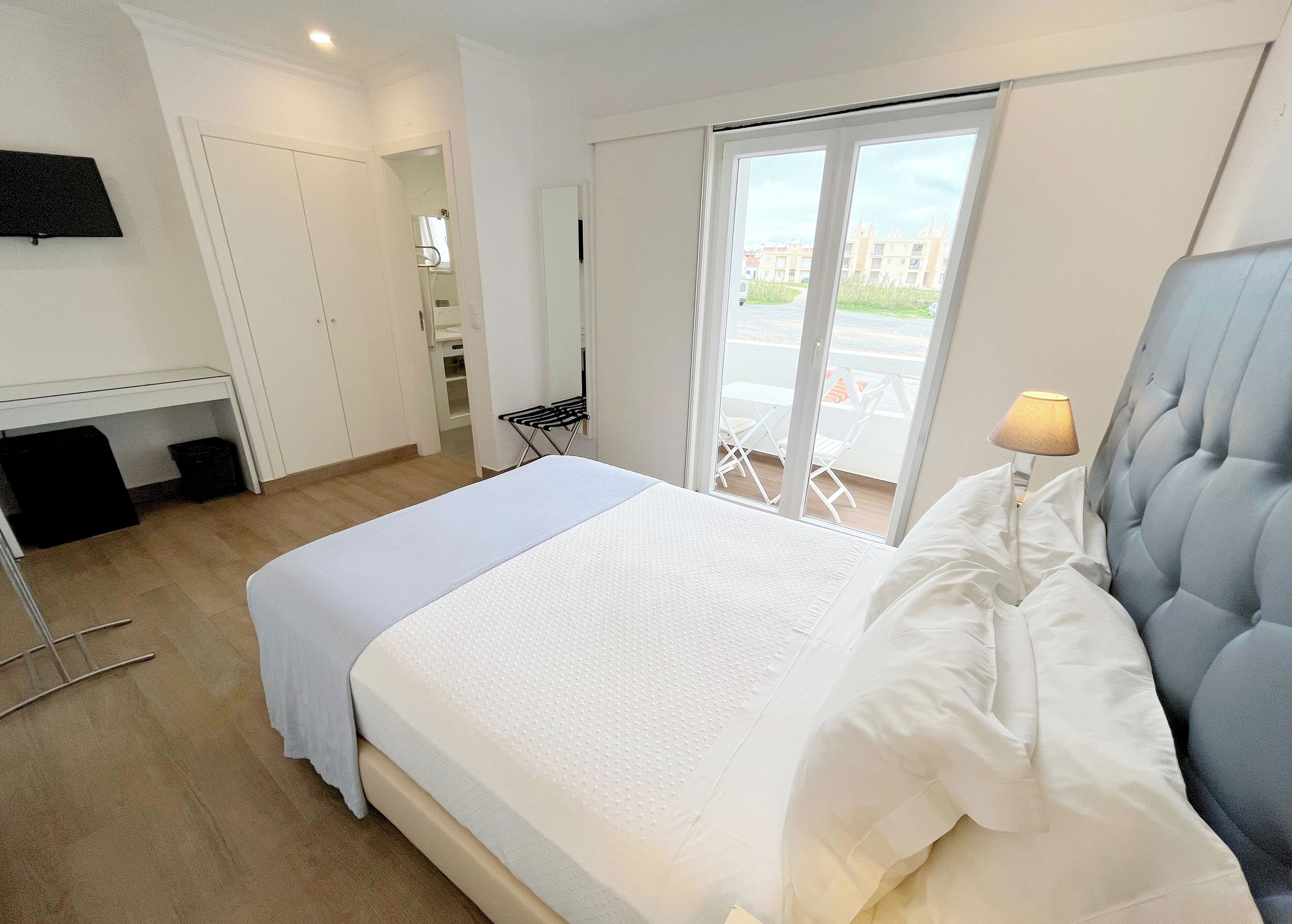 A hotel room with a bed, a desk, a chair, a TV, and a window with a view of buildings.
