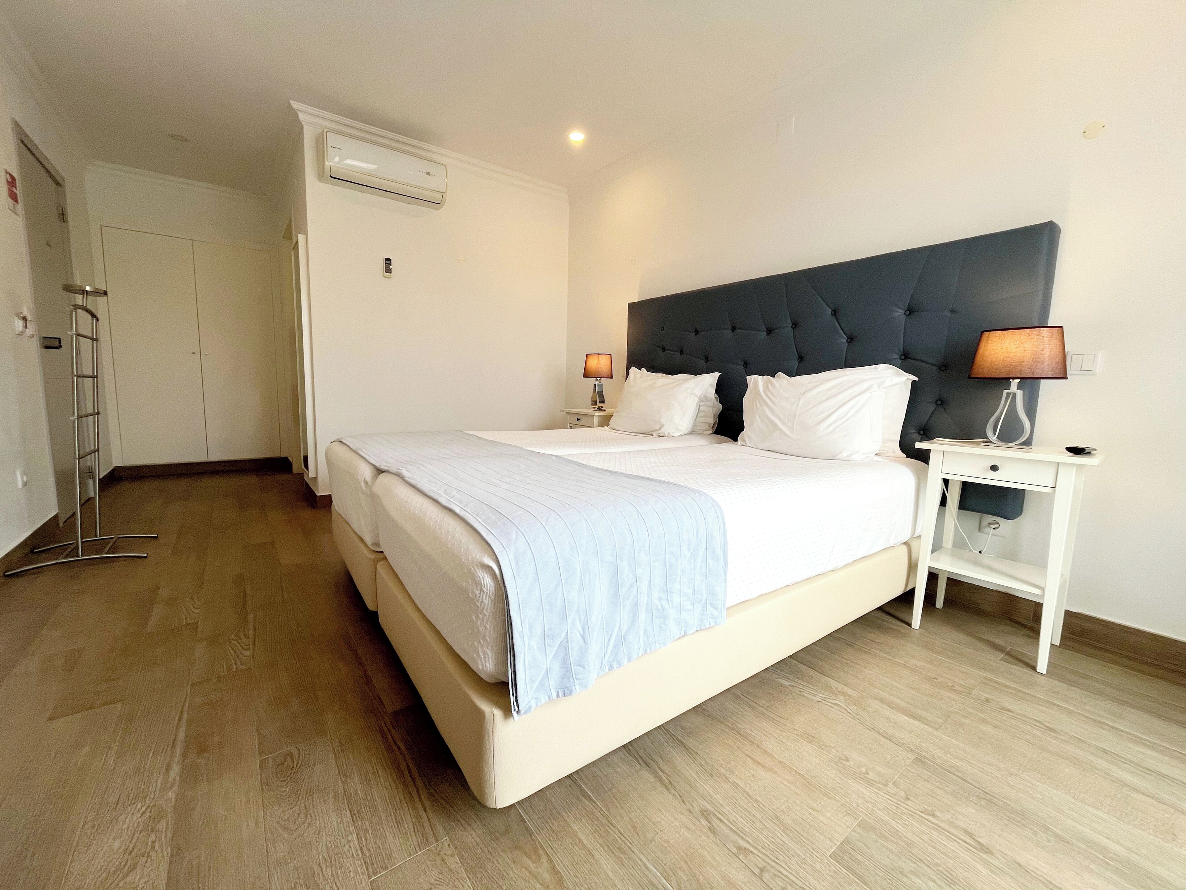 A neatly made bed with a headboard, a bedside table with a lamp, and an air conditioning unit on the wall.