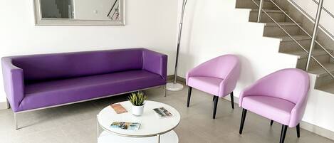 Una sala de estar moderna con un sofá morado, dos sillones rosas y una mesa de centro blanca con revistas.
