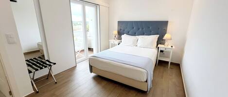 A modern bedroom with a large bed, a sliding glass door to a balcony, and a small table with a lamp.