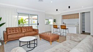 A modern living room with a brown leather sofa, a white round coffee table, and a grey sofa. There are two wooden chairs and a small ottoman. A kitchen area is visible in the background with wooden cabinets and a white countertop. - Freedom Beach Homes Unit 199B Brand New in Rainbow Bay with private plunge pool and Wi-Fi Inc. (Coolangatta)