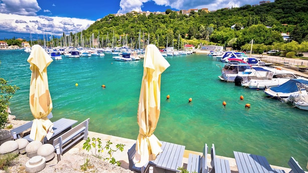 A marina with boats, a wooden deck with chairs and umbrellas, and a hillside with buildings in the background. - Holiday apartment with terrace and sea view (Njivice)