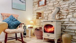 A cozy room with a stone wall, a wooden chair with a blue cushion, a white lamp, and a wood-burning stove. - 2 bedroom accommodation in Acton (Acton)