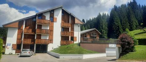 A multi-story building with wooden balconies, a parking area, and a mountainous backdrop.