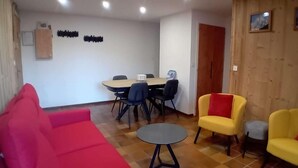 Una habitación con un sofá rojo, sillones amarillos, una mesita redonda, una mesa de comedor con sillas y paneles de madera en las paredes.