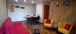 Um quarto com um sofá vermelho, poltronas amarelas, uma mesa redonda pequena, uma mesa de jantar com cadeiras e painéis de madeira nas paredes.