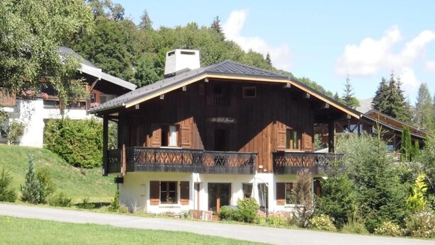 Una gran cabaña de madera con balcón, rodeada de exuberante vegetación y un cielo despejado.