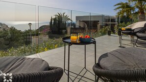 Un balcon doté d'une barrière en verre, de meubles en osier, d'une table avec un panier de fruits et des boissons, et offrant une vue sur l'océan et la végétation.