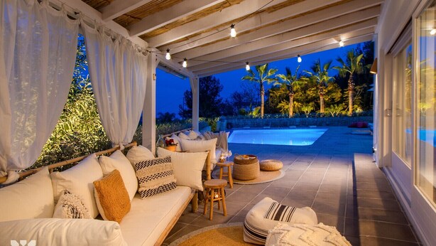 Un área de estar al aire libre con techo, piscina, asientos acolchados e iluminación ambiental.