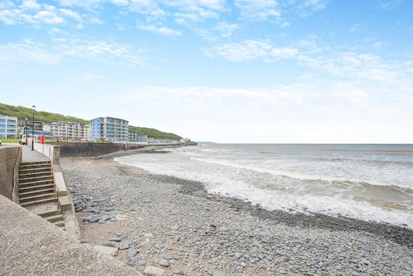 A coastal hotel with a pebble beach, a staircase leading to the beach, and buildings with balconies overlooking the sea.