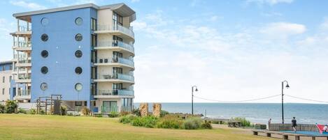 Un immeuble à plusieurs étages doté de balcons, avec un espace verdoyant à l’avant et une vue sur la mer.