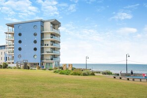 Un immeuble à plusieurs étages doté de balcons, avec un espace verdoyant à l’avant et une vue sur la mer.