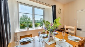 Een eetkamer met een tafel gedekt voor vier personen, een raam met uitzicht op groen en een klok aan de muur.