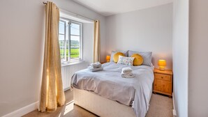 Een slaapkamer met een groot raam, een bed met kussens, een nachtkastje met een lamp en uitzicht op groen buiten.