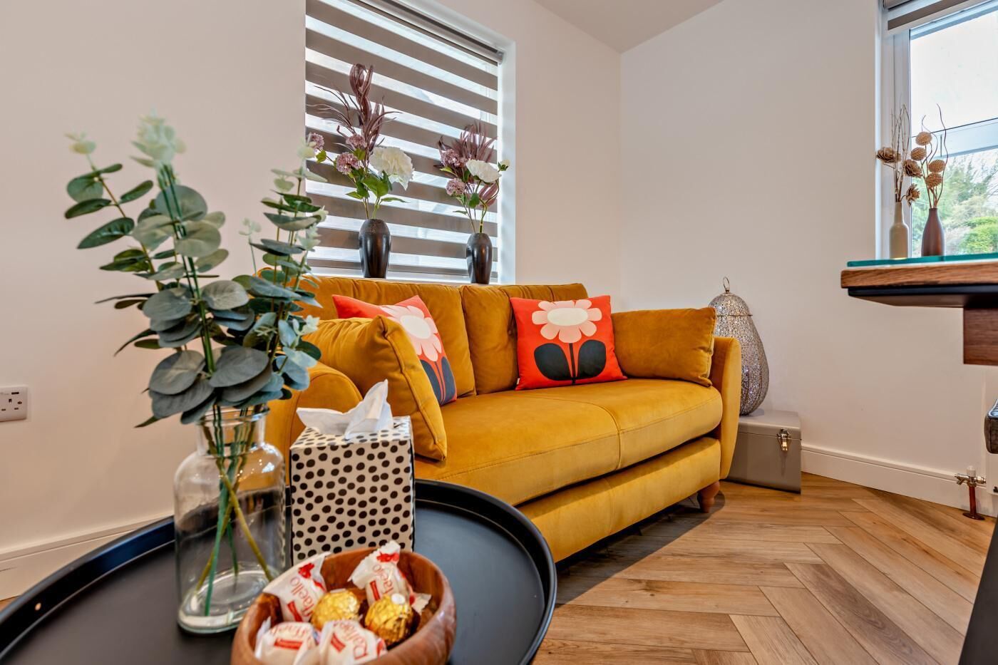 Ein Wohnzimmer mit einem senfgelben Sofa, einem runden Couchtisch und einer Vase mit Grünpflanzen.