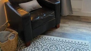 Un sillón de cuero negro con un cojín, ubicado junto a una ventana con cortinas grises, una alfombra estampada en el suelo y una cesta a la izquierda.