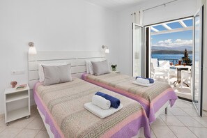 Un dormitorio con dos camas, una mesita de noche con lámpara y vista al mar a través de un ventanal.
