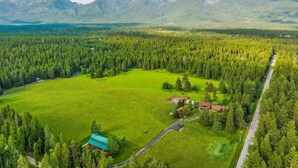 An aerial view of a property with a large green field, a small building with a red roof, and a road curving through the landscape. - G-Bar-N Ranch Pendleton Log Home (Bigfork)