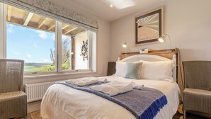 Une chambre à coucher avec un grand lit, deux fauteuils, une fenêtre donnant sur un beau paysage et une tête de lit ornée d’une roue décorative.
