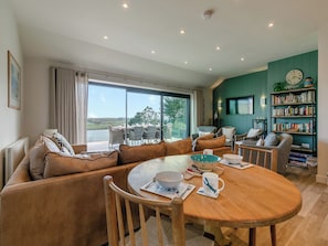 A modern living room with a large window, a round wooden dining table, leather sofas, a bookshelf, and a television mounted on the wall.