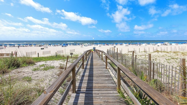Ein hölzerner Steg führt zu einem Sandstrand mit Sonnenschirmen und Badegästen.