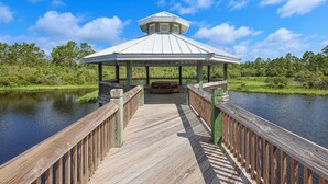 Ein hölzerner Steg führt zu einem Gazebo mit einem Metalldach, umgeben von Wasser und viel Grün.