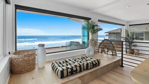 Una sala de estar moderna con un ventanal grande con vistas al océano, una cesta de mimbre, un jarrón blanco y un banco estampado.
