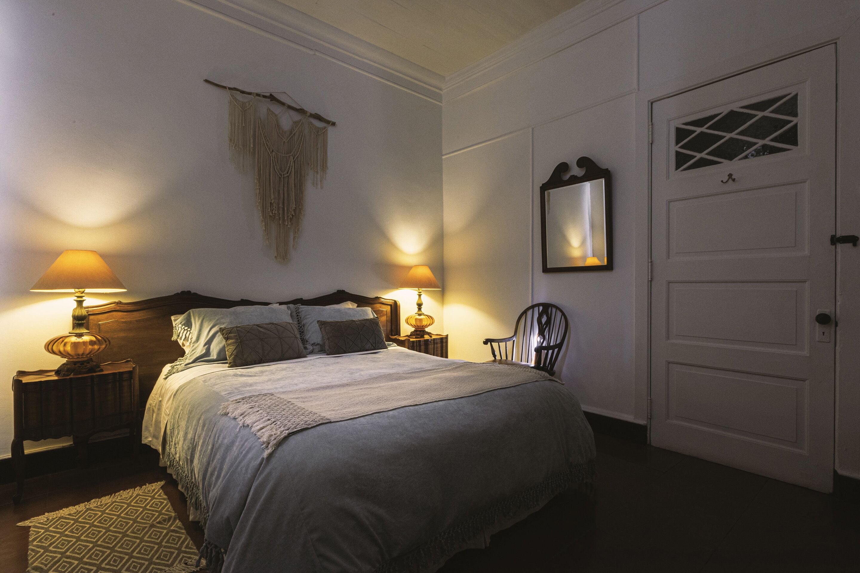 A neatly made bed with a headboard, two bedside lamps, a decorative wall hanging, and a door with a window.