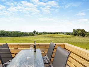 A table with chairs and a bottle of wine, set on a wooden deck with a view of a green field and trees.