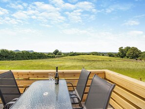 A table with chairs and a bottle of wine, set on a wooden deck with a view of a green field and trees. - Rabbits Rest - UK46128 (Nesscliffe)