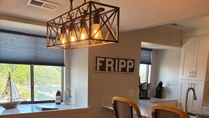 Een eetgedeelte met een houten tafel, geweven placemats, rieten stoelen, een hanglamp en een bord aan de muur.