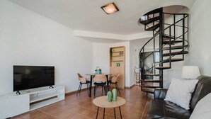A modern living room with a flat-screen TV, a leather sofa, a dining area with chairs, a small table with a cactus, and a spiral staircase. - Santa Barbara Serena Apartment Jávea (Javea / Xàbia)