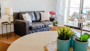 A living room with a dark leather sofa, a round coffee table, and a lamp on a side table. There are potted plants and a vase with flowers on the coffee table. - Santa Barbara Serena Apartment Jávea (Javea / Xàbia)