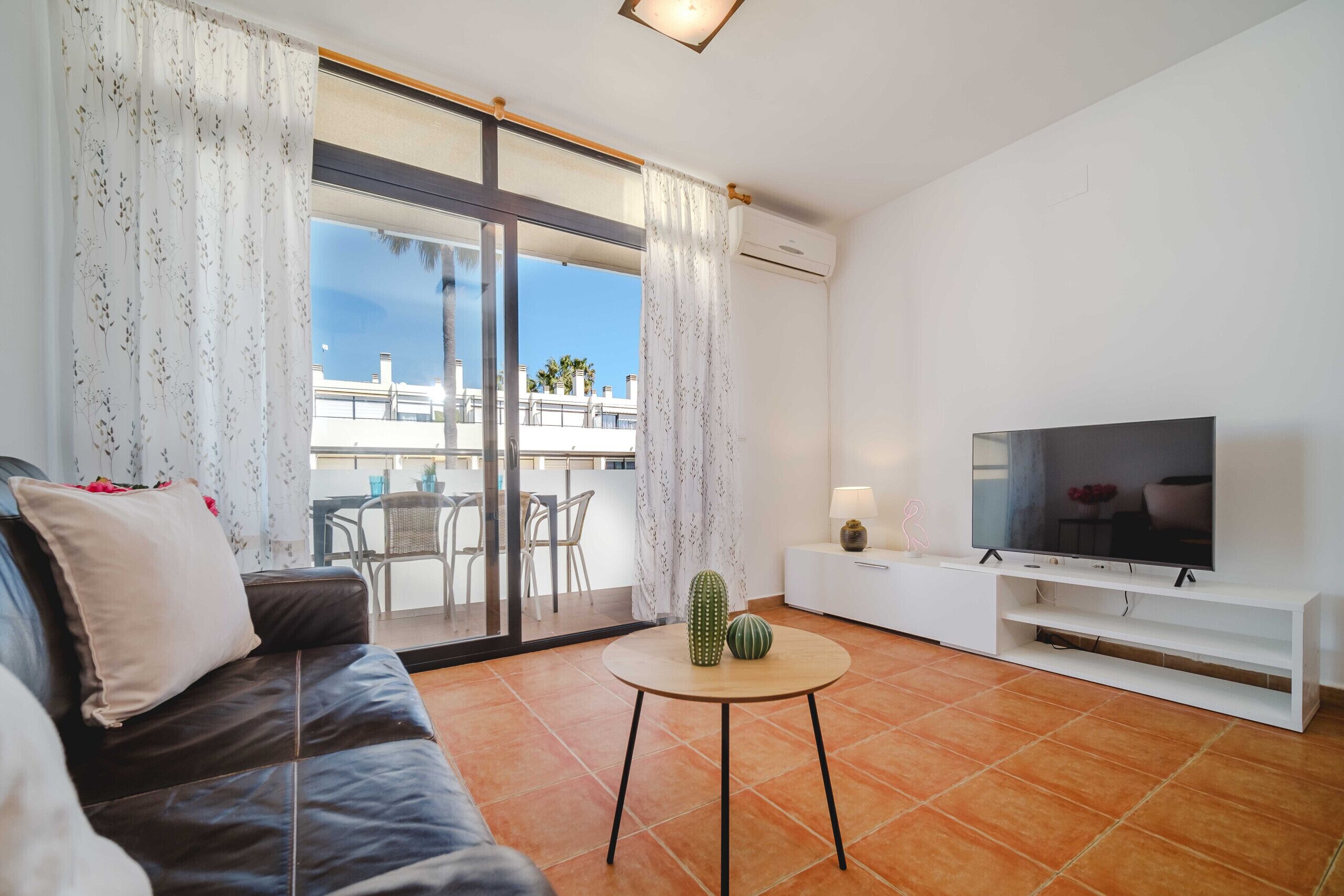 A modern living room with a leather sofa, a flat-screen TV, a small round table with decorative items, and a sliding glass door leading to an outdoor balcony.