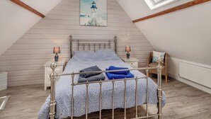 Ein Schlafzimmer mit schrägem Dach, Dachfenstern, einem Bett mit gemusterten Betttextilien, Nachttischlampen und einem Gemälde eines Leuchtturms.