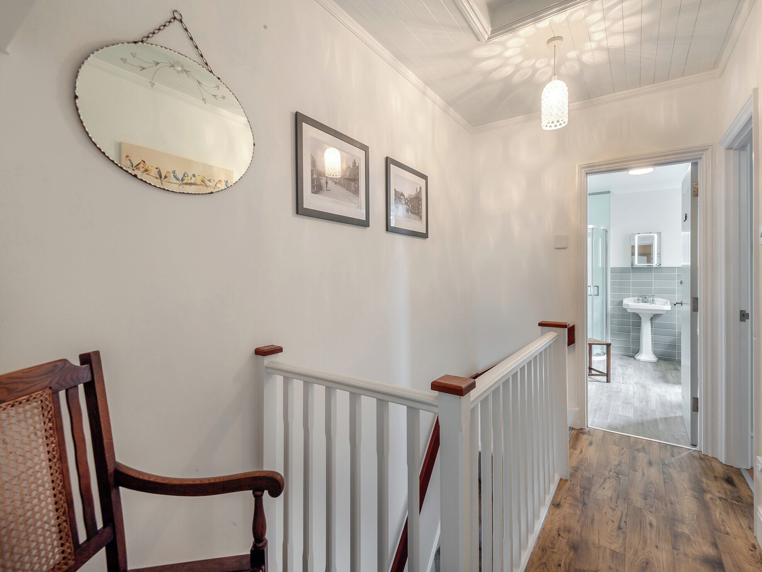 Un couloir avec une chaise en bois, un radiateur blanc et un miroir suspendu. Des tableaux encadrés ornent le mur, et un luminaire est fixé au plafond.