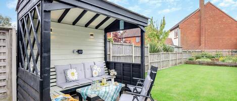 A covered outdoor seating area with a table and chairs, a bench, and a garden view.