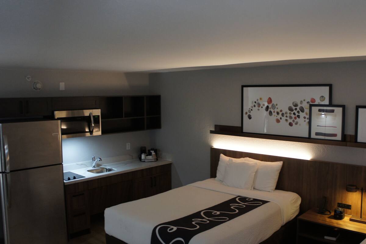 A hotel room with a bed, a nightstand, a bedside table, a microwave, a refrigerator, a sink, and framed artwork on the wall.